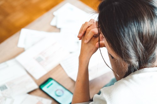 Image shows a woman looking at a number of bills and invoices with her phone set to a banking app.